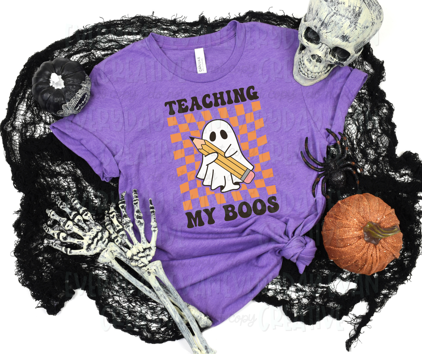 A flat lay of a t-shirt surrounded by Halloween-themed items. The shirt's design includes an orange checker design with a sheet ghost on top holding a large pencil. Text above and below the design, in block font, reads, "teaching my boos."