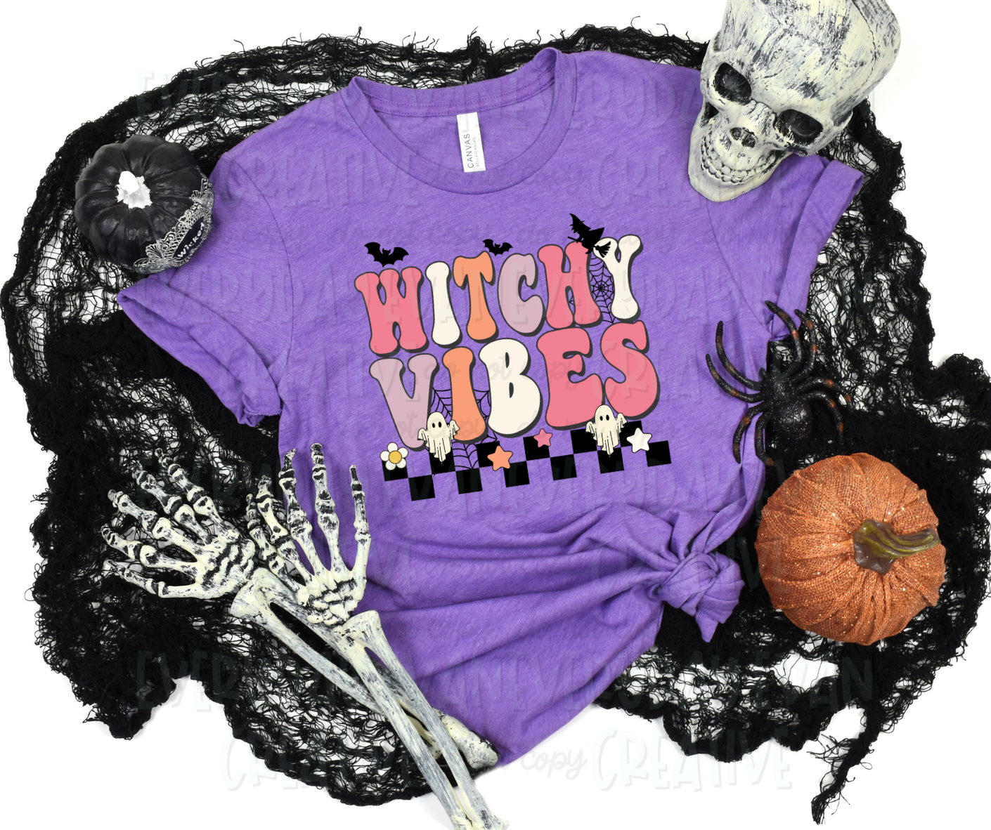 Image displays a t-shirt surrounded by Halloween-themed items. The shirt's design includes funky block font text that reads, "witchy vibes." The words sit above a strip of black checkered design and has webs, sheet ghosts, bats, and star surrounding the design.