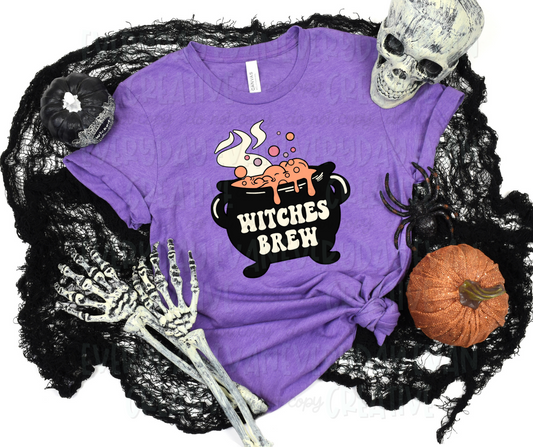 Image displays a t-shirt surrounded by Halloween-themed items. The shirt's design includes a black cauldron with "bitches brew" printed in block letters on it. The cauldron has orange bubbling liquid and steam rising from it.