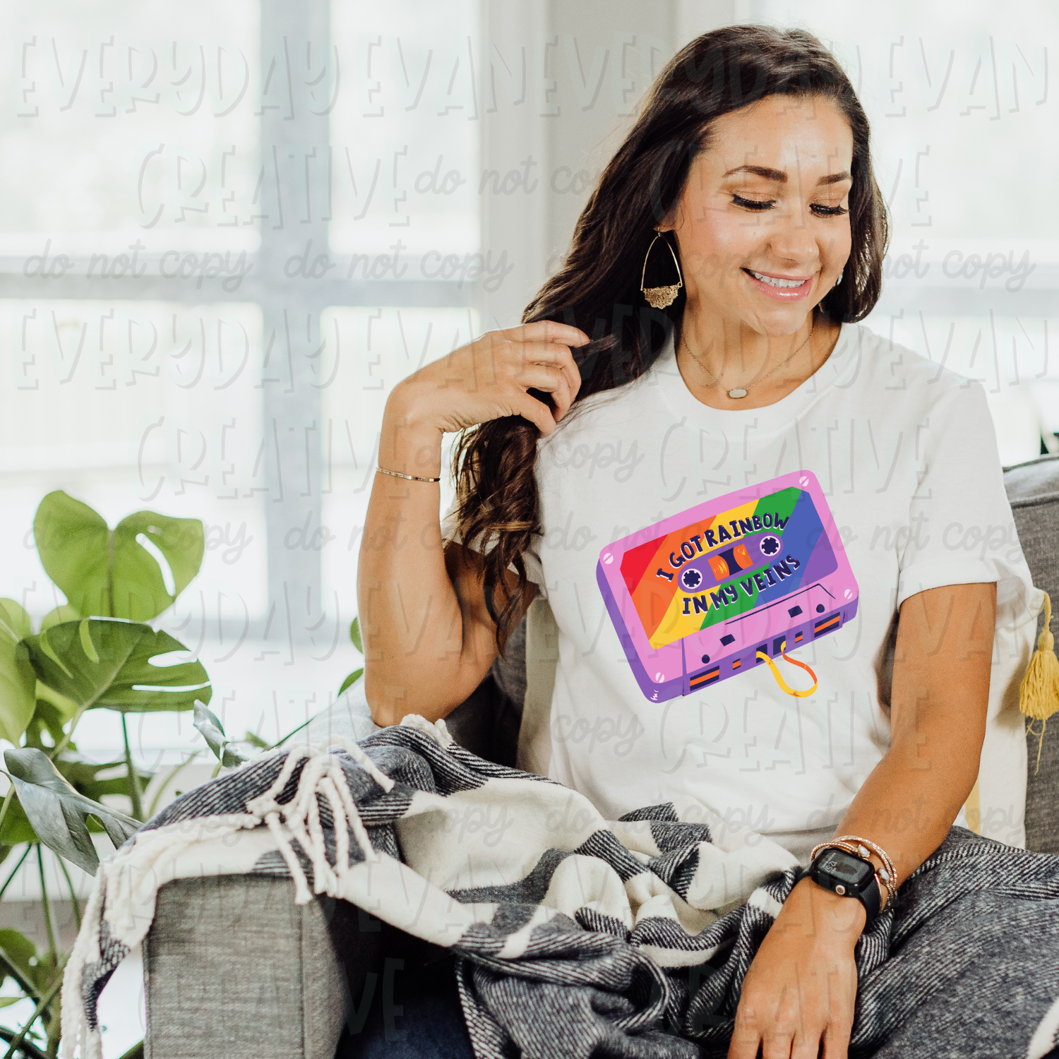A brunette white woman sits on a couch modeling a white t-shirt. The design on the t-shirt is a retro illustration of a cassette tape. The base of the tape is purple and the inner part where a label would be is rainbow striped and in blue capital letters it says "I Got Rainbow In My Veins."