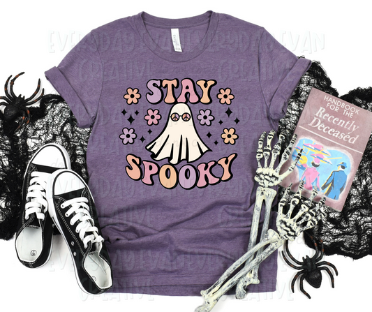 A flat lay of a t-shirt surrounded by Halloween-themed and household items. The shirt's design includes funky, multicolored pastel lettering that reads, "stay spooky" above and below a sheet ghost wearing peace symbol shades. Flowers and starbursts surround the design.