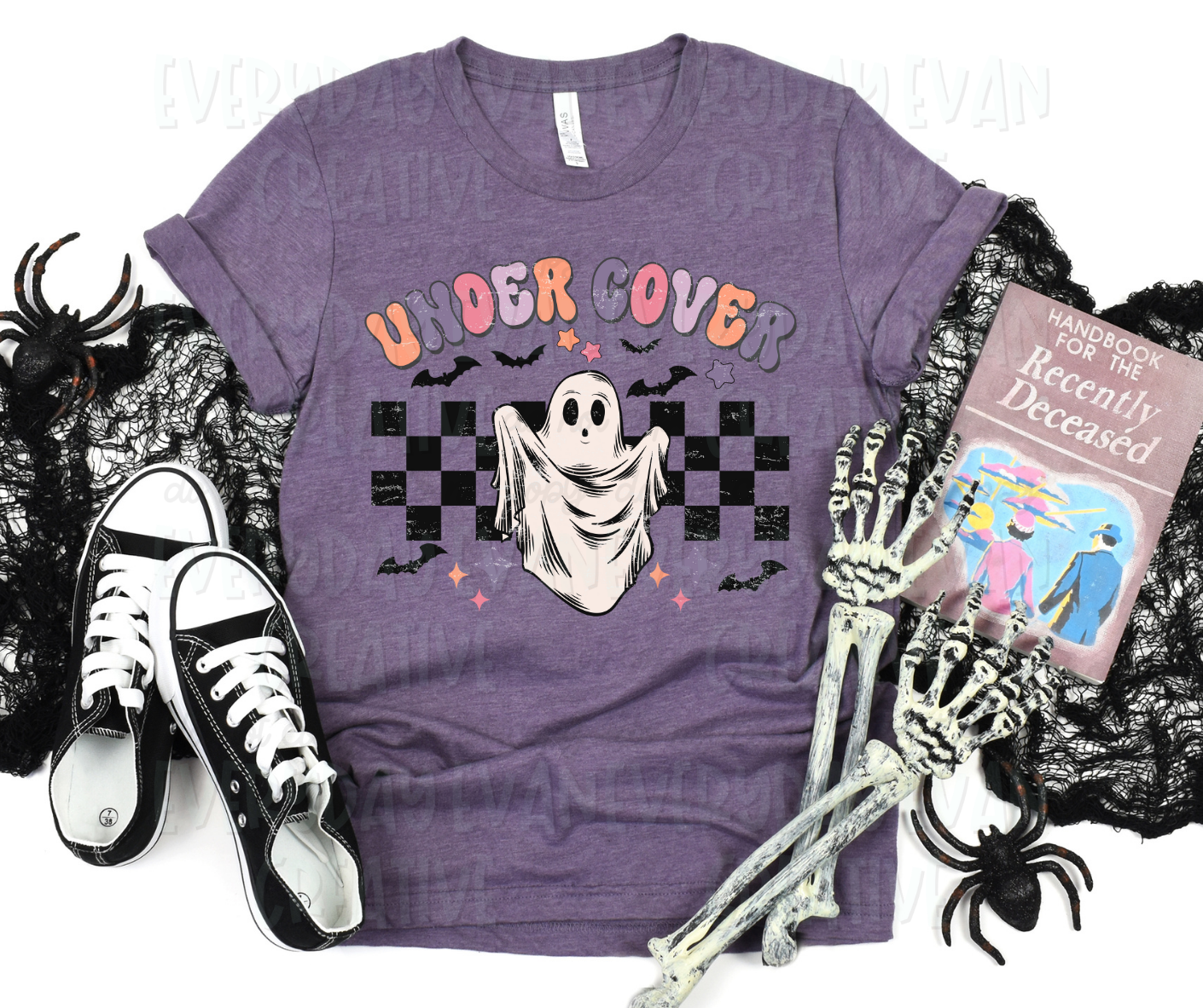 A flat lay of a t-shirt surrounded by miscellaneous Halloween-themed items. The shirt's design has a checkered background behind a faced sheet ghost. Stars, starbursts, and bats surround the design. Above the design, in funky block font text, it reads, "under cover."
