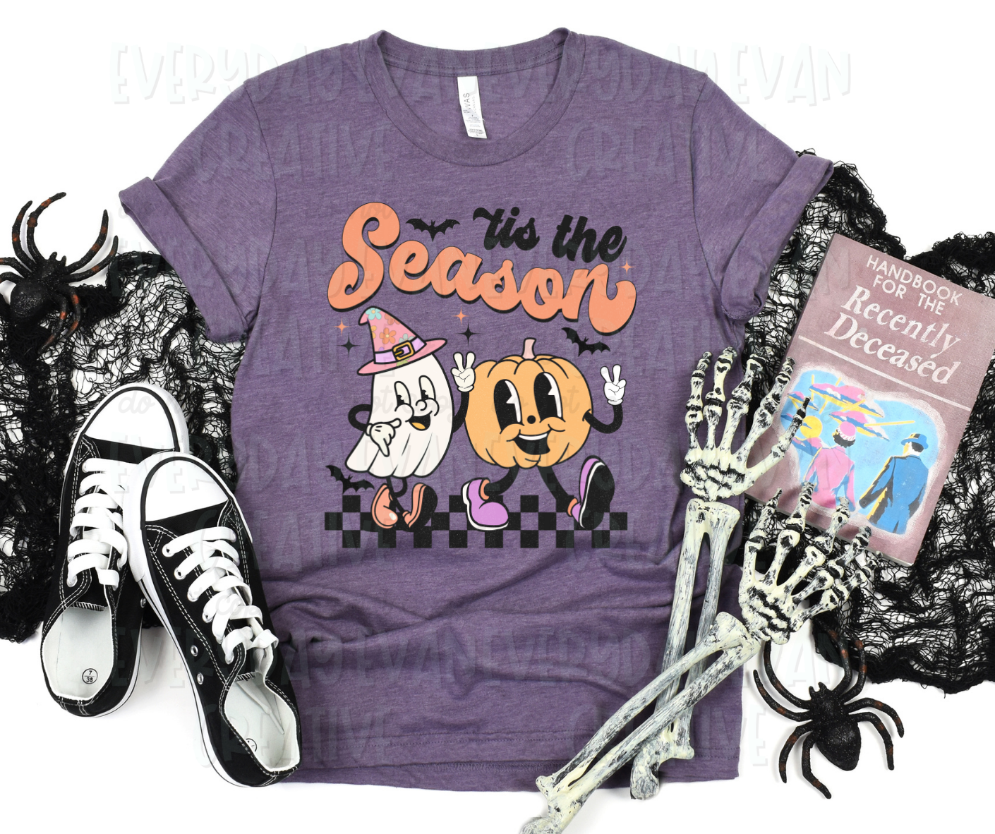 A flat lay of a t-shirt surrounded by miscellaneous Halloween-themed items. The shirt's design includes a personified sheet ghost wearing a witch hat and a personified pumpkin walking beside the ghost. They both extend peace signs with their hands and walk down a checkered print. Starbursts and bats surround the design decoratively. Above the design, in large script font, it reads, "tis the season."