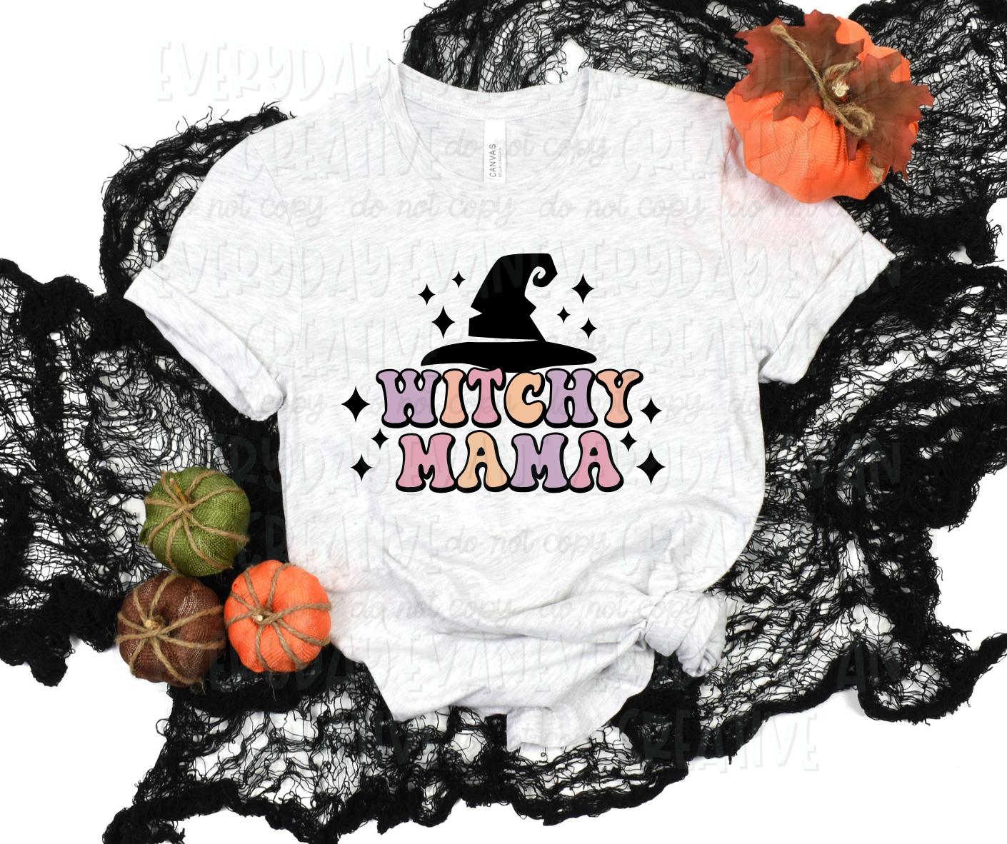 A flat lay of a t-shirt surrounded by fall-themed decorations. The shirt's design includes funky block font text that reads, "witchy mama" in alternating pale purple, orange, and pink colors. Above the text, there is a large black witch hat. Black starbursts surround the design.