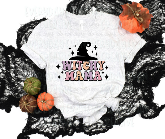 A flat lay of a t-shirt surrounded by fall-themed decorations. The shirt's design includes funky block font text that reads, "witchy mama" in alternating pale purple, orange, and pink colors. Above the text, there is a large black witch hat. Black starbursts surround the design.