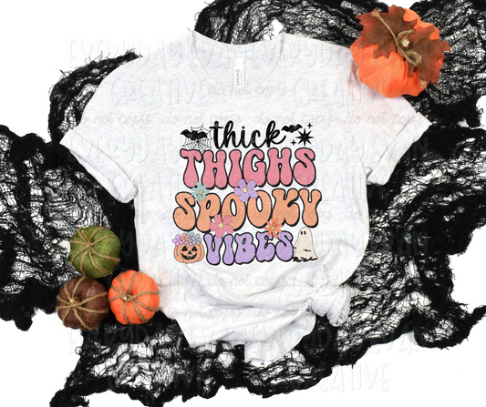 Image displays a t-shirt surrounded by fall-themed decorations. The shirt's design includes text, in funky bubble font and script font, that reads, "thick thighs spooky vibes." A pumpkin, sheet ghost, starbursts, flowers of various sizes and colors, bats, and a spider web accent the design. 