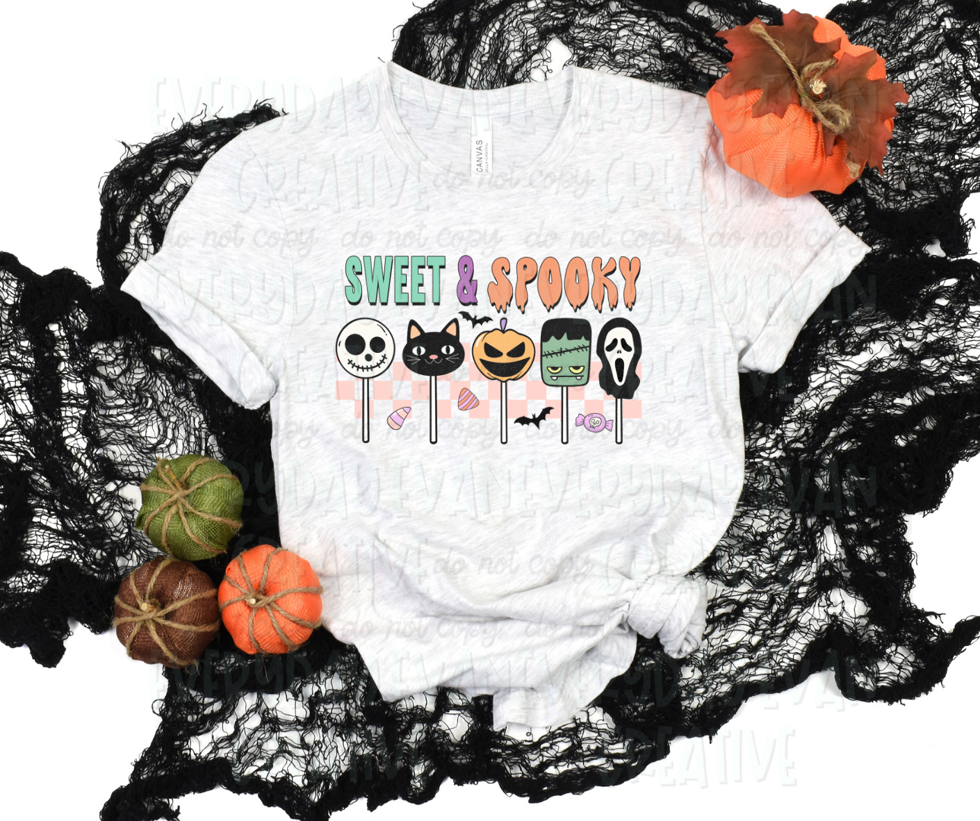 A flat lay of a t-shirt surrounded by fall-themed decorations. The shirt's design includes text that reads "sweet and spooky" in various fonts and Halloween colors. Below the text, there are several cake pops designed as Halloween figures including a cartoon skeleton, a black cat, a faced pumpkin, a cartoon Frankenstein, and a Scream mask. They are above a pink checkered design and surrounded by candies and bats. 
