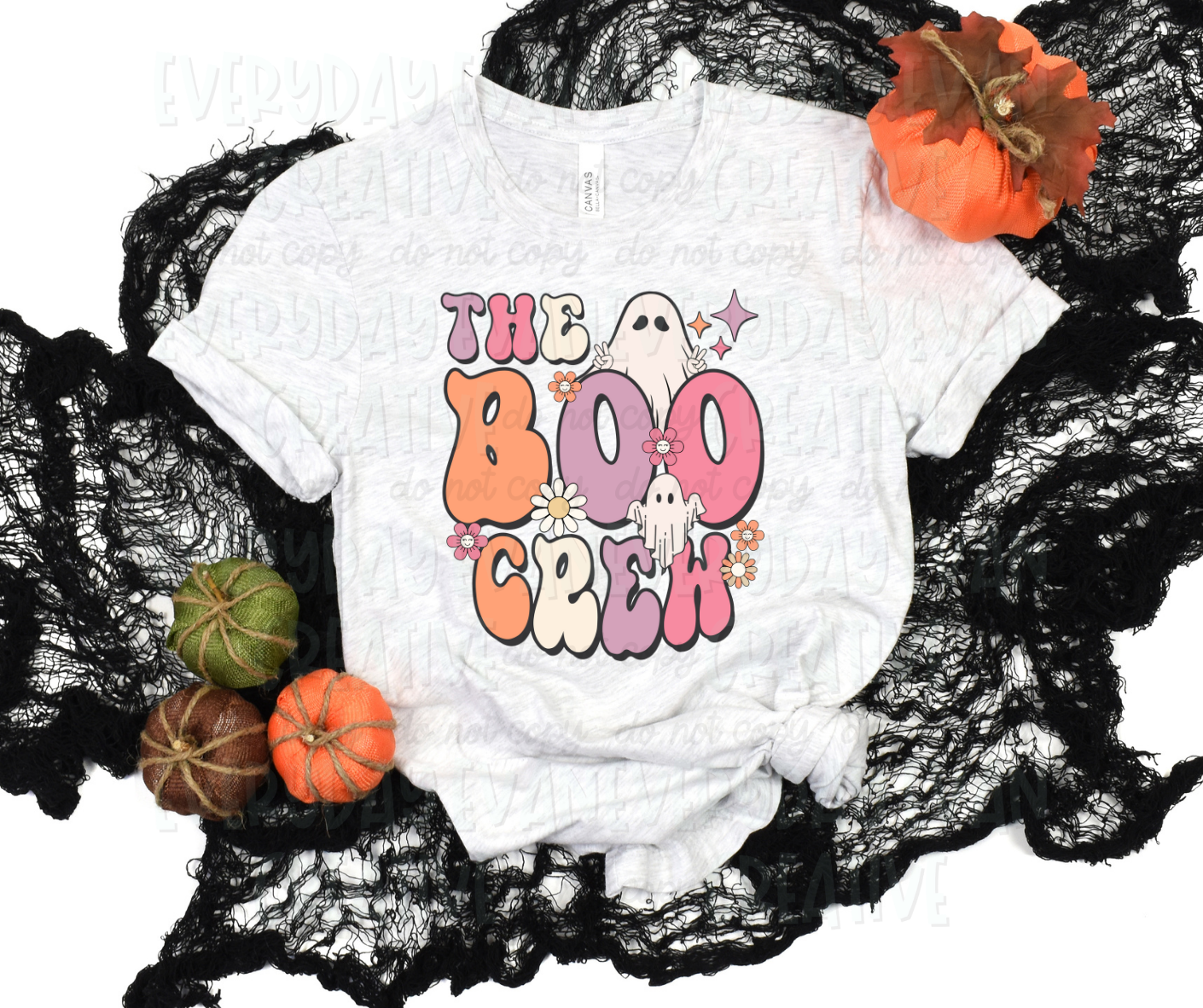 A flat lay of a t-shirt surrounded by fall-themed decorations. The shirt's design includes text, in script bubble font, that reads, "the boo crew" in alternative pale purple, pink, and white colors. Personified sheet ghosts, flowers of various types, colors, and sizes, and starbursts surround the design.