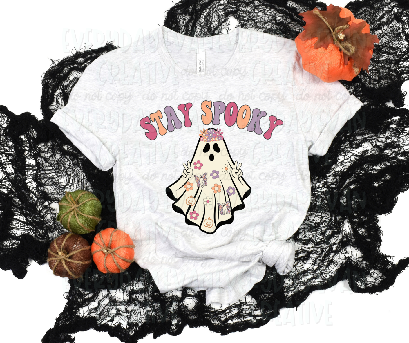 Image displays a t-shirt surrounded by fall-themed decorations. The shirt's design includes a sheet ghost with pale yellow, purple, pink, and white flowers, butterfly ghosts, and pumpkins on it. It also wears a flower crown and is holding up peace signs. Above the design, in funky block font, it reads, "Stay spooky" in alternative pink, yellow, and purple.