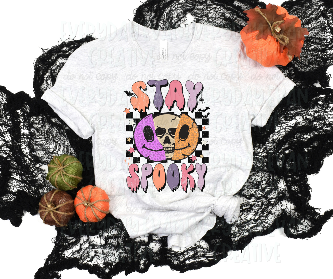 A flat lay of a t-shirt surrounded by fall-themed decorations. The shirt's design includes a black and white checkered print behind a split-in-half smiling face with a skull inside. The smiling face is half purple and half orange. Different colored stars, black bats, and a spider accent the design. Above and below the design, in funky block font that drips, it reads, "stay spooky" in alternating orange, purple, pink, and light purple.