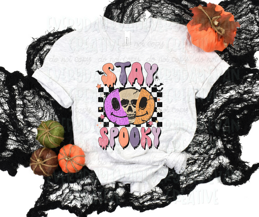 A flat lay of a t-shirt surrounded by fall-themed decorations. The shirt's design includes a black and white checkered print behind a split-in-half smiling face with a skull inside. The smiling face is half purple and half orange. Different colored stars, black bats, and a spider accent the design. Above and below the design, in funky block font that drips, it reads, "stay spooky" in alternating orange, purple, pink, and light purple.