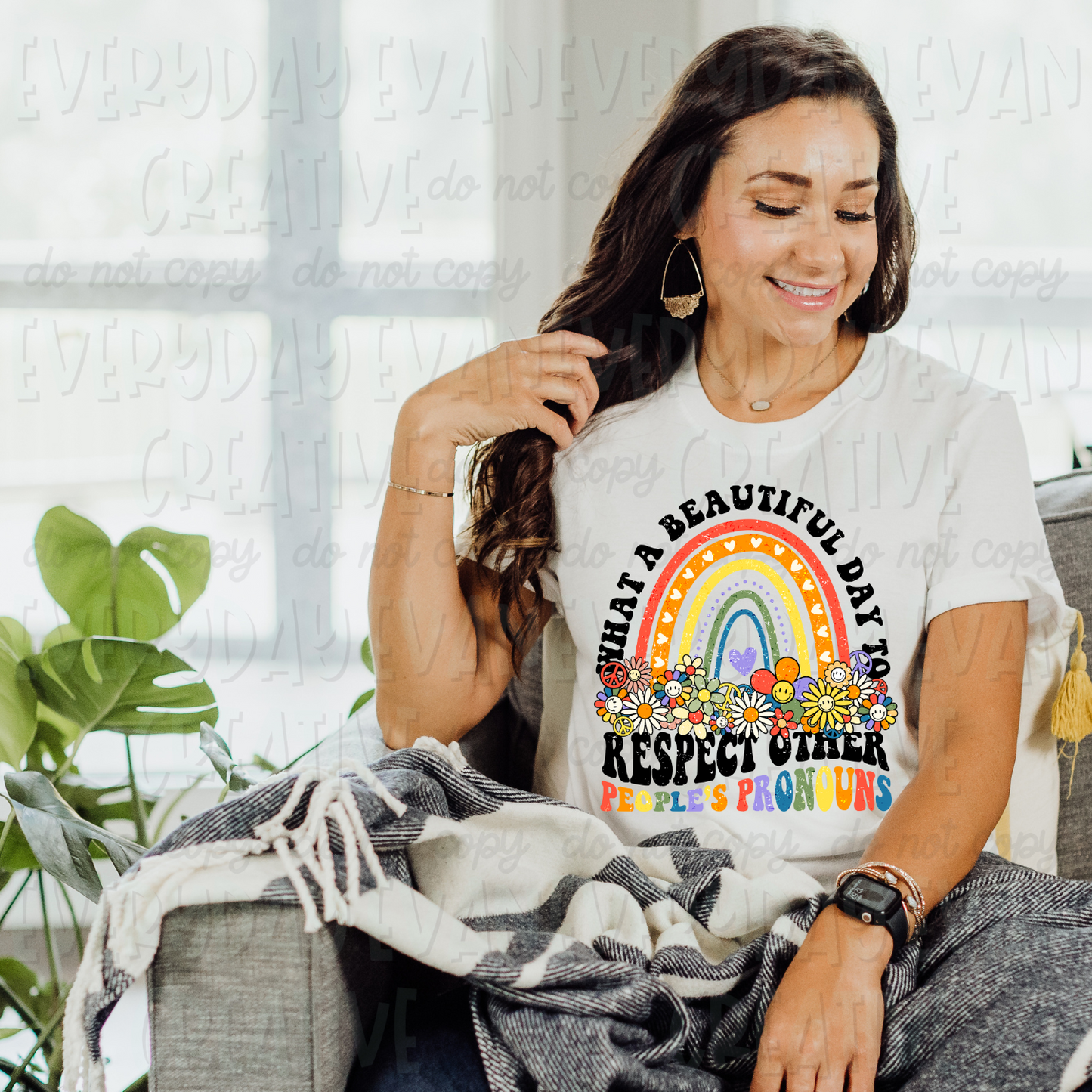 A brunette white woman is sitting on a couch modeling a white t-shirt. The design on the shirt is groovy/retro in nature. It has a brightly colored rainbow in the center with groovy flowers at its base. Above the rainbow in black retro font it says "what a beautiful day" and underneath the rainbow and flowers it says in black wavy retro font "respect other." Underneath in rainbow wavy retro font it says, "people's pronouns."