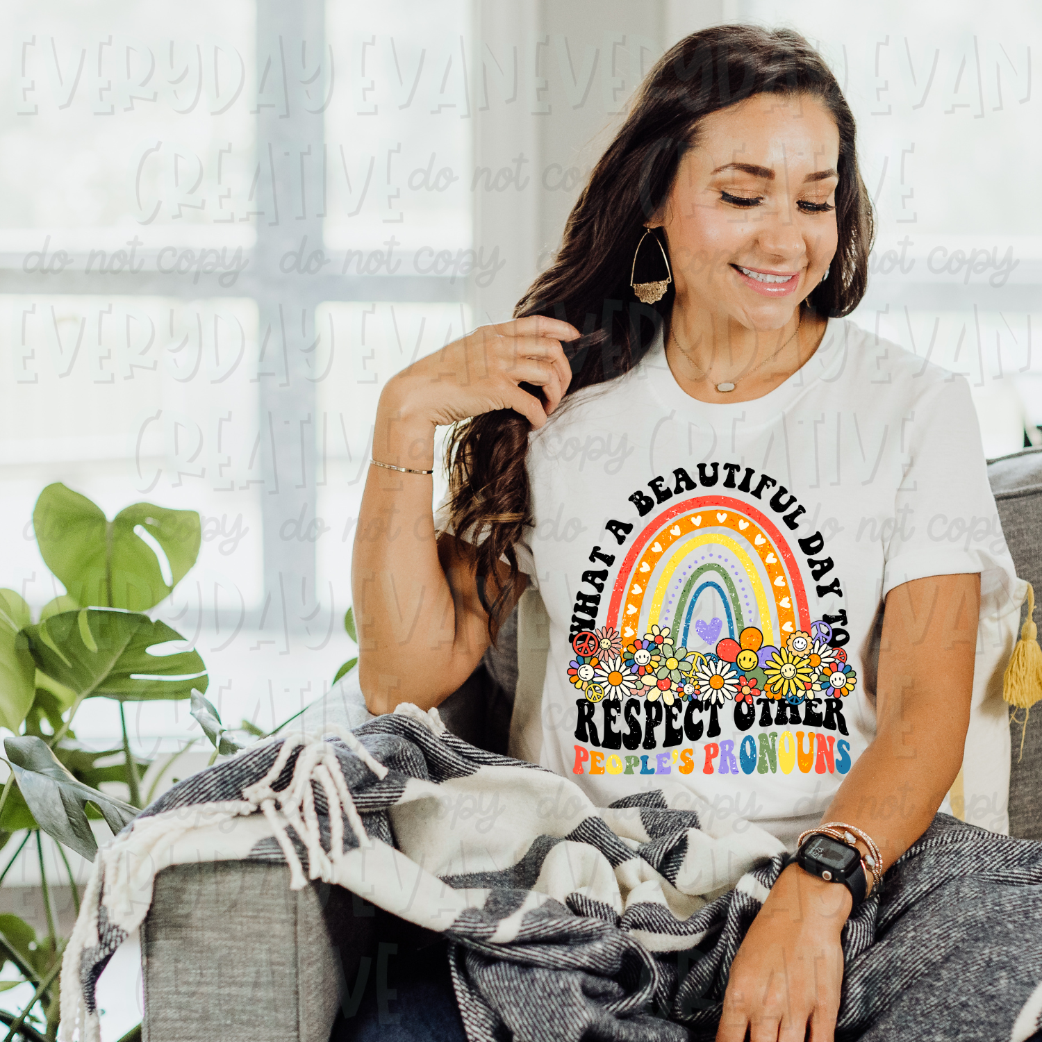 A brunette white woman is sitting on a couch modeling a white t-shirt. The design on the shirt is groovy/retro in nature. It has a brightly colored rainbow in the center with groovy flowers at its base. Above the rainbow in black retro font it says "what a beautiful day" and underneath the rainbow and flowers it says in black wavy retro font "respect other." Underneath in rainbow wavy retro font it says, "people's pronouns."