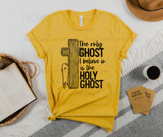 Images displays a yellow t-shirt with books and other generic accessories surrounding it. The shirt has black script and block text font that reads "The only ghost I believe in is the holy ghost" on one half. "Ghost" and "holy ghost" are in block font, and "the only" and "I believe in is the" is in script font. The other half of the design, flush with the text, has a large wooden cross with a ghost peeking out from behind it. 
