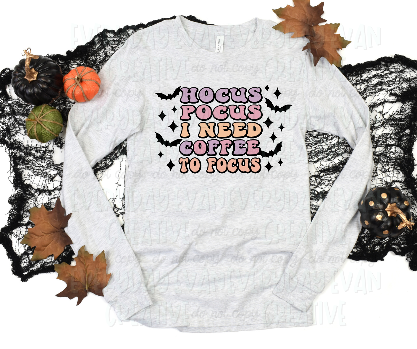 Image displays a long sleeved shirt surrounded by fall-themed decorations. The shirt's design includes text that reads, "hocus pocus I need coffee to focus" in bubbled font and alternative pale purple, pink, and orange colors. The text is surrounded by black bats.  