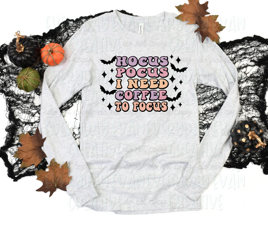 Image displays a long sleeved shirt surrounded by fall-themed decorations. The shirt's design includes text that reads, "hocus pocus I need coffee to focus" in bubbled font and alternative pale purple, pink, and orange colors. The text is surrounded by black bats.  