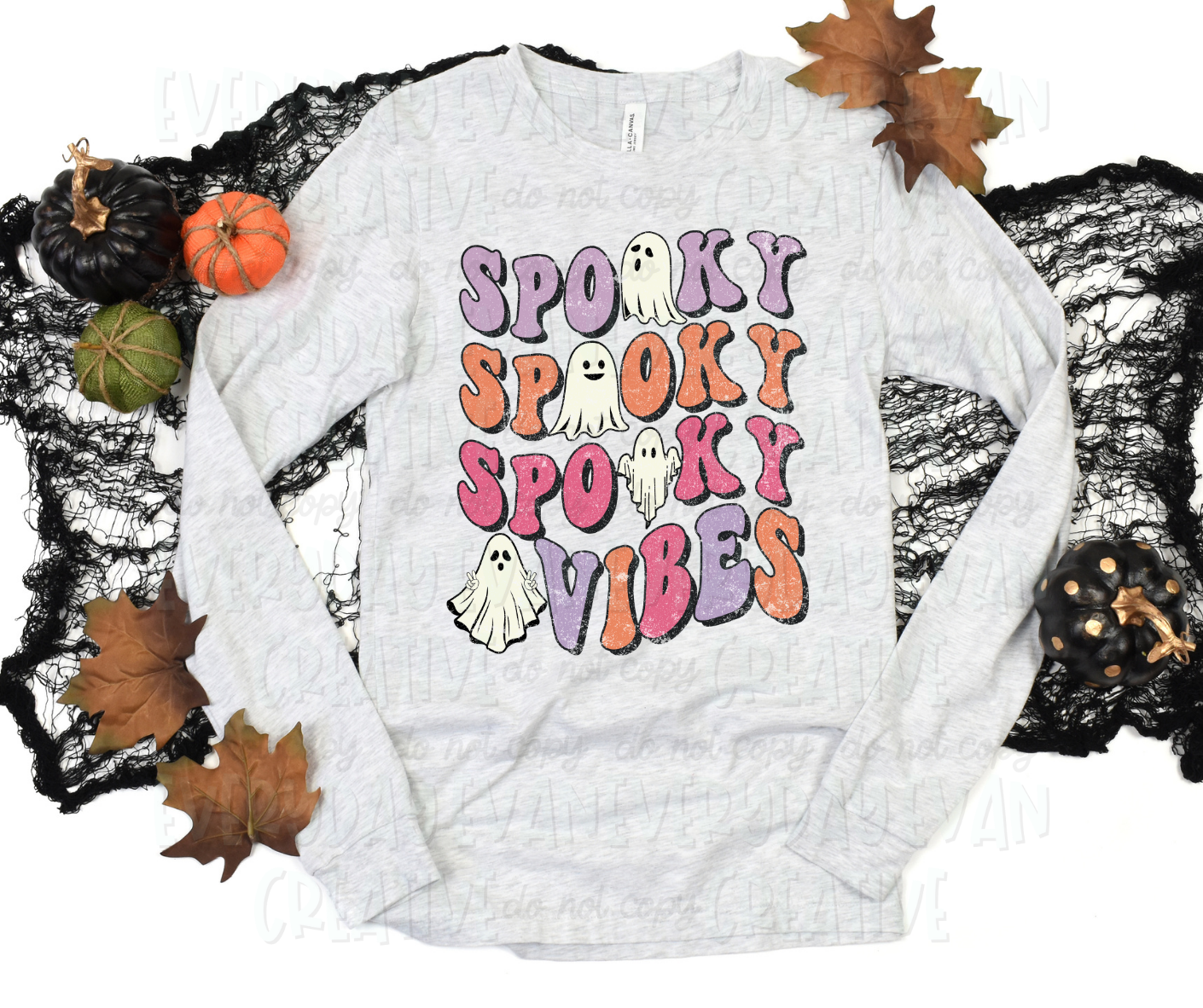 A flat lay of a t-shirt surrounded by fall-themed decorations. The shirt's design includes funky, block test with a shadow effect that reads, "spooky spooky spooky vibes." Several o's are sheet ghosts on the design, and several of them have faces.