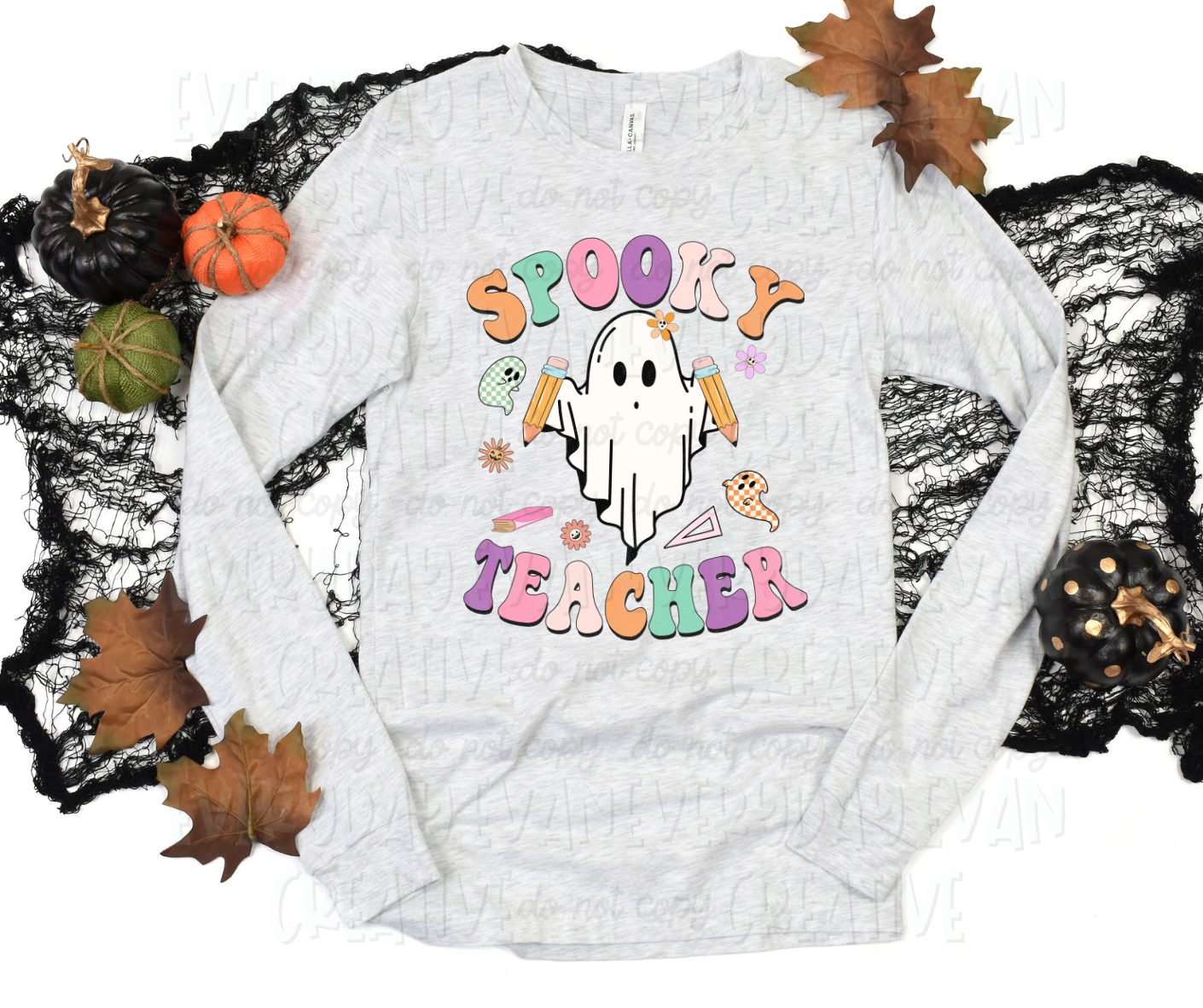 A flat lay of a t-shirt surrounded by fall-themed decorations. The shirt's design includes a personified sheet ghost holding pencils surrounded by miscellaneous school items and other ghosts in checkered font. Text surrounding the design, in funky block text of alternating orange, green, pink, purple, and light pink, reads "spooky teacher."