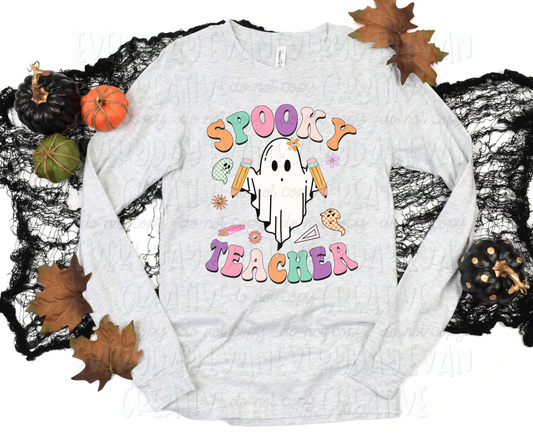 A flat lay of a t-shirt surrounded by fall-themed decorations. The shirt's design includes a personified sheet ghost holding pencils surrounded by miscellaneous school items and other ghosts in checkered font. Text surrounding the design, in funky block text of alternating orange, green, pink, purple, and light pink, reads "spooky teacher."