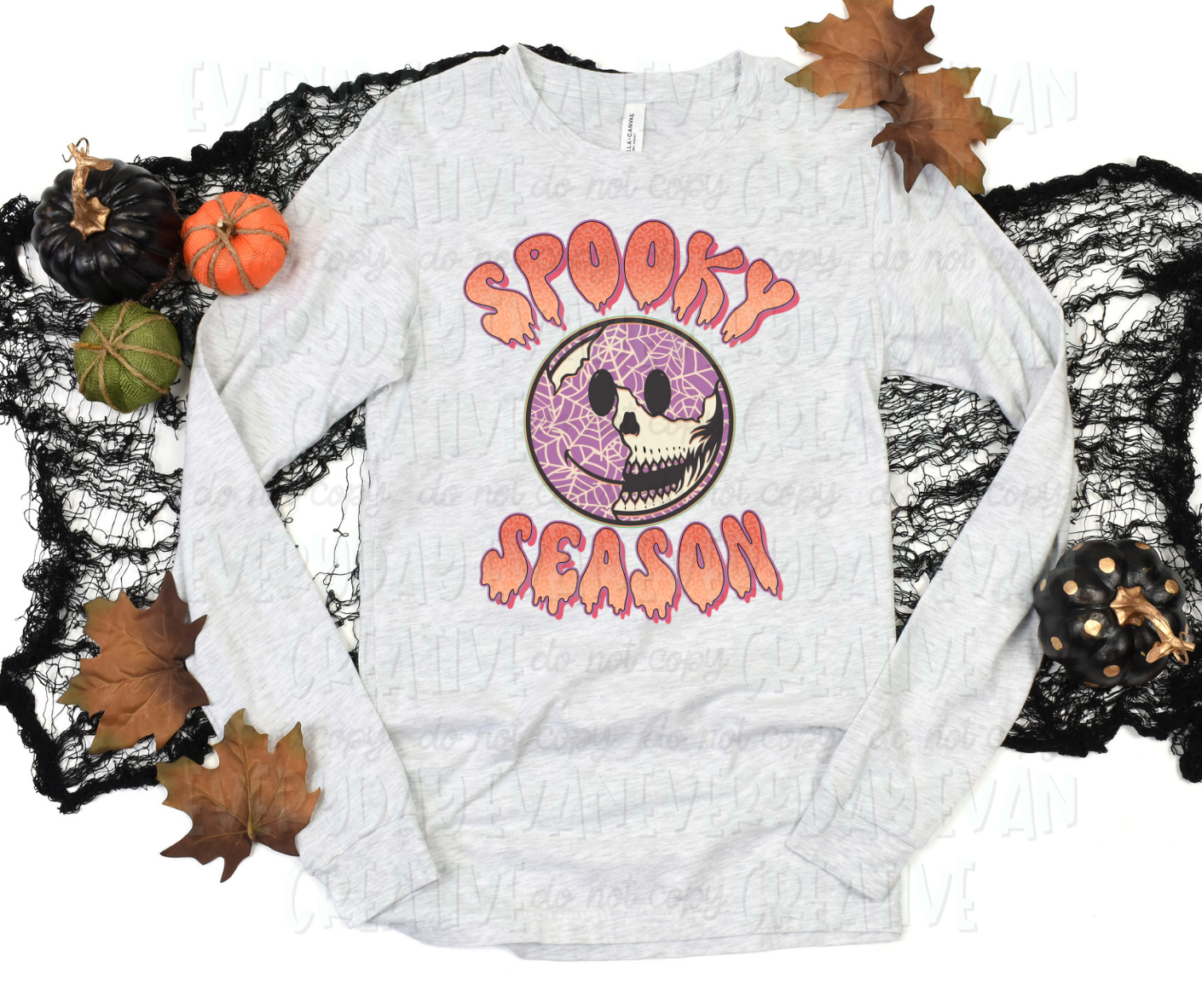 Image displays a t-shirt surrounded by fall-themed decorations. The shirt's design includes Halloween-themed font text with a dripping effect that reads "spooky season." The text surrounds a smiling face with a crackling effect exposing a skull underneath. The smiling face that is intact is purple with webbing on it.