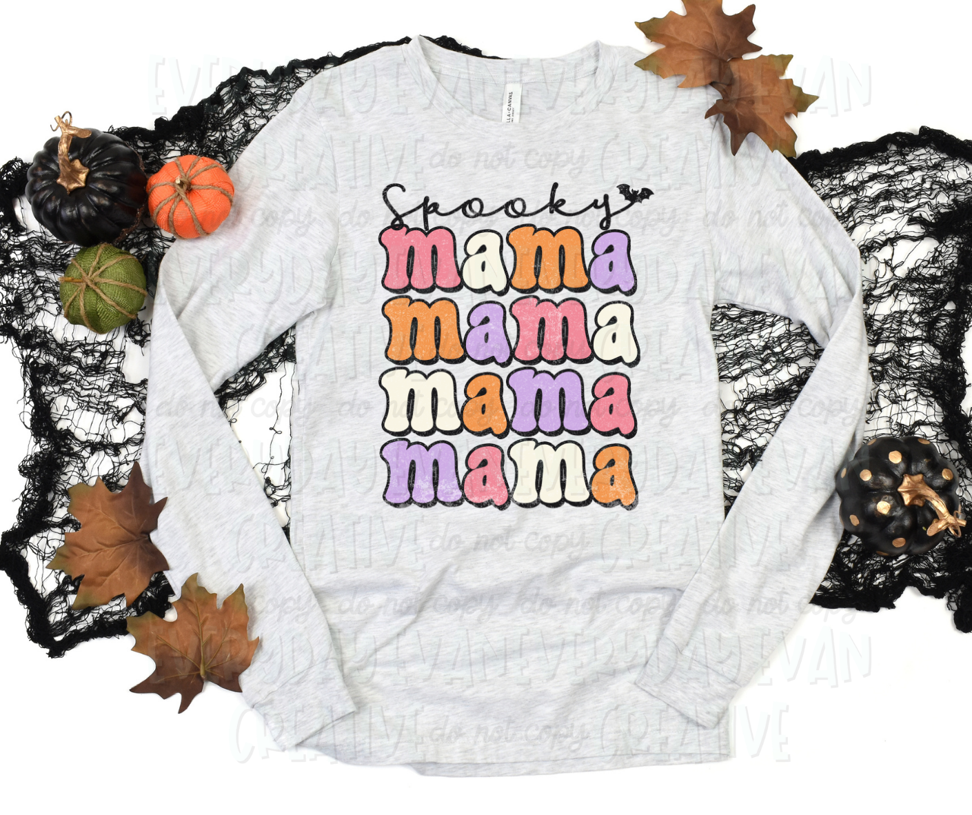 A flat lay of a t-shirt surrounded by fall-themed decorations. The shirt's design includes text that reads, "spooky mama mama mama mama" in script and block text. "Spooky" is written in cursive script and ends with a bat symbol. The "mama" text is in funky block text with alternating pink, white, orange, and purple colors.