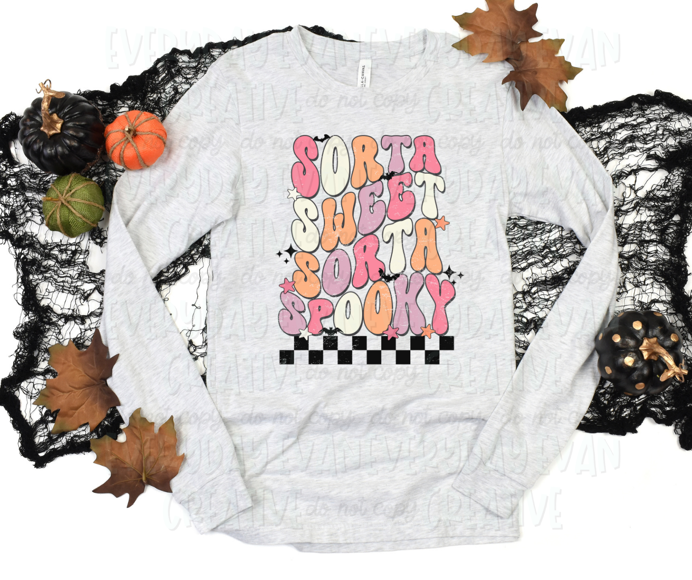 A flat lay of a t-shirt surrounded by fall-themed decorations. The shirt's design includes text, in funky block font, that reads, "sorta sweet sorta spooky" in alternating pink, orange, purple, and white colors. The text is above a checkered design. The design is surrounded by starbursts, stars, and bats.