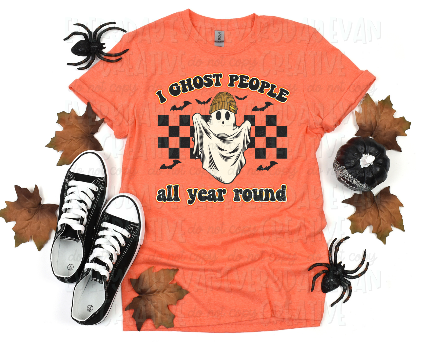 A flat lay of a t-shirt surrounded by fall-themed decorations. The shirt's design includes a checkered design behind a personified sheet ghost in a beanie. Bats surround the design. Above and below the design, text in block font reads, "I ghost people all year round."