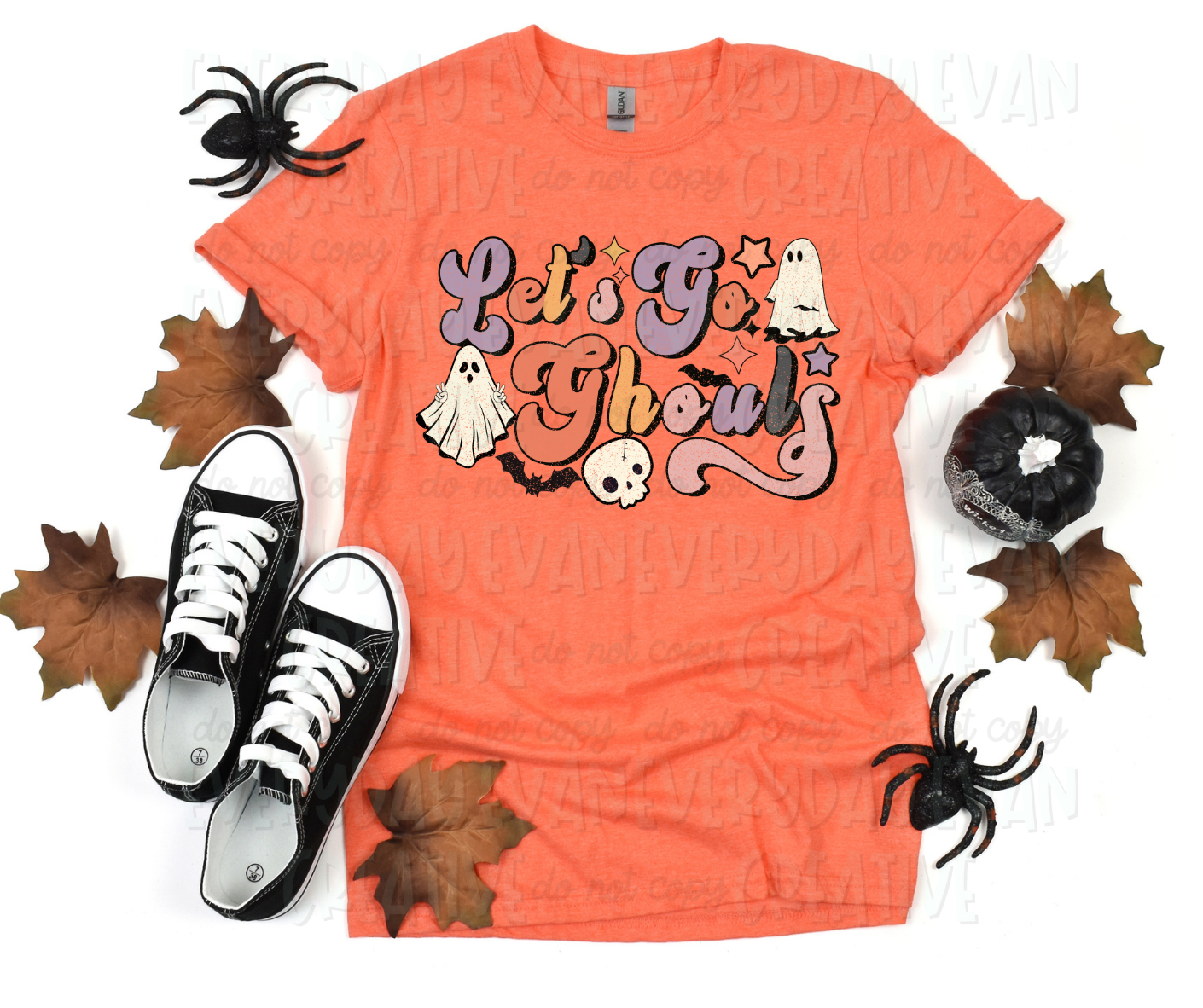 A flat lay of a t-shirt surrounded by fall-themed decorations. The shirt's design includes text, in script block text, that reads, "let's go ghouls" in alternaiting purple, orange, yellow, black, and light pink colors. Personified sheet ghosts, starbursts, a cartoon skull, and bats surround the design.