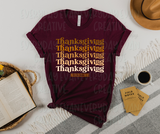 Image displays a burgundy colored shirt surrounded by miscellaneous desk items. The shirt has script text that reads "thanksgiving" five times up and down the shirt. The words have a gradient from orange-brown to white. Under the text, there is a orange box with "1 Thessalonians 5:18" written inside in the same color as the shirt. 