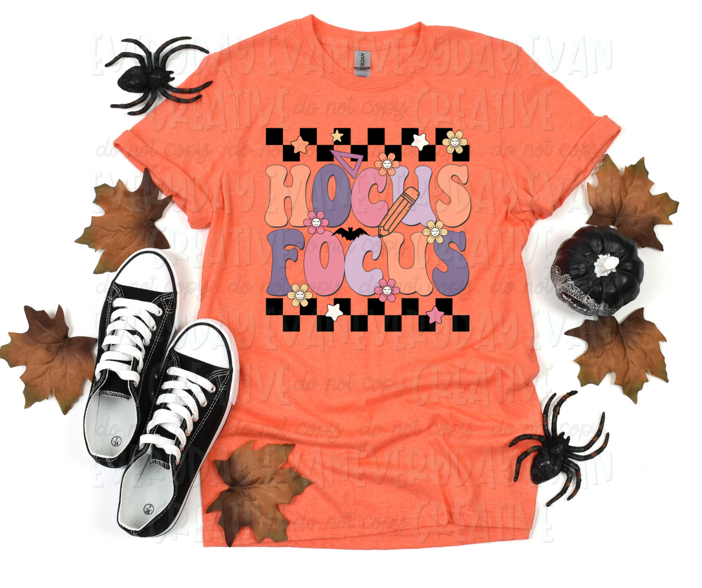 Image displays a t-shirt surrounded by fall-themed decorations. The shirt's image includes text, in funky block font, that reads "hocus focus" in alternating orange, purple, and pink colors. The text is in between checkered strips with flowers, a bat, school supplies, and stars surrounding the design.