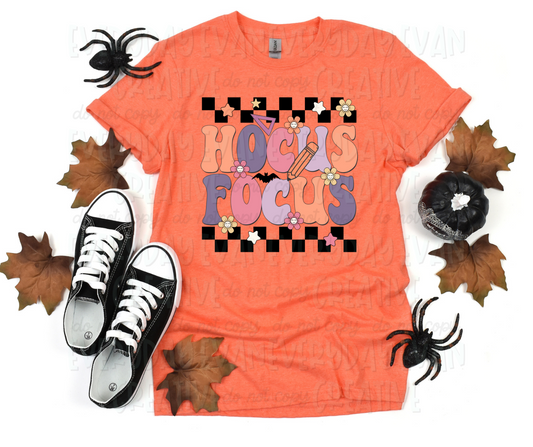 Image displays a t-shirt surrounded by fall-themed decorations. The shirt's image includes text, in funky block font, that reads "hocus focus" in alternating orange, purple, and pink colors. The text is in between checkered strips with flowers, a bat, school supplies, and stars surrounding the design.