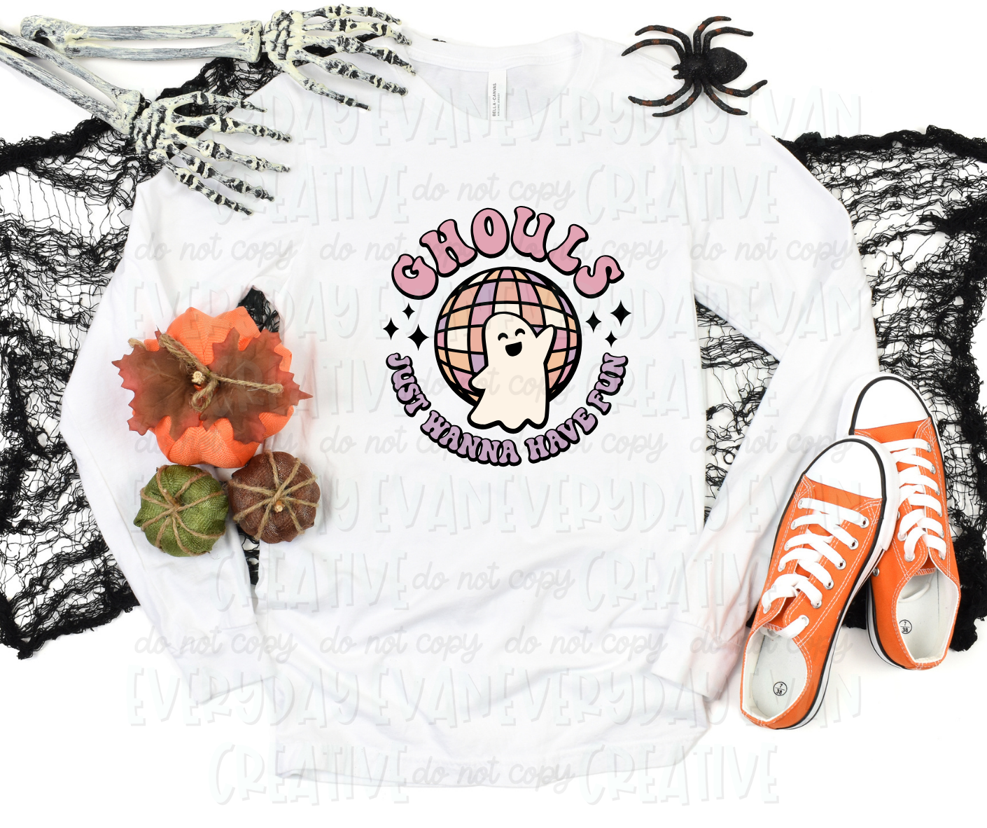 A flat lay of a t-shirt surrounded by Halloween-themed decorations. The shirt's design includes a personified ghost dancing in front of a colorful disco ball. Text surrounding the design in funk block font reads, "ghouls just wanna have fun" separated by starbursts.