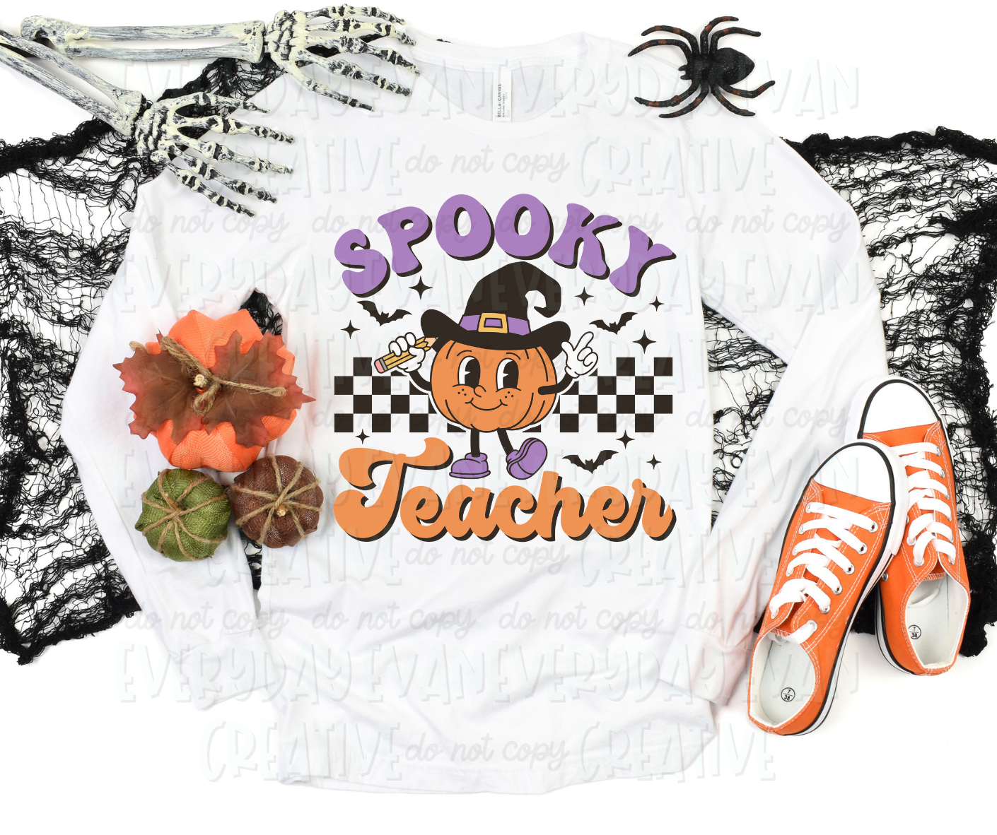 A flat lay of a t-shirt surrounded by Halloween-themed decorations. The shirt's design includes a personified pumpkin holding a pencil and wearing a witch hat. The pumpkin is in front of a checkered design surrounded by bats and starbursts. Text surrounding the design reads, "spooky teacher" in a script block font in orange and purple colors.