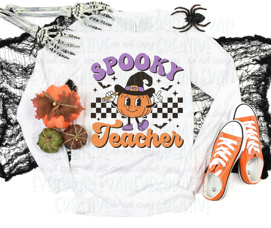 A flat lay of a t-shirt surrounded by Halloween-themed decorations. The shirt's design includes a personified pumpkin holding a pencil and wearing a witch hat. The pumpkin is in front of a checkered design surrounded by bats and starbursts. Text surrounding the design reads, "spooky teacher" in a script block font in orange and purple colors.