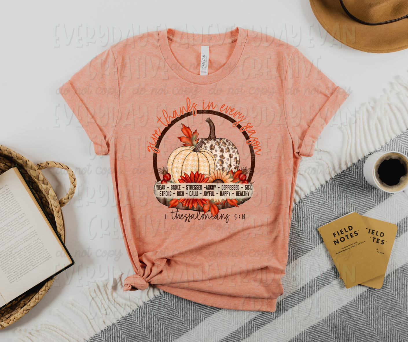 Image displays a t-shirt surrounded by miscellaneous desk items. The shirt's image includes a circle with decorative pumpkins and fall foliage inside. The bottom of the circle has a white box with different emotions listed inside in two rows reading, "weak, broke, stressed, angry, depressed, sick, strong, rich, calm, joyful, happy, healthy." The top of the circle has script text reading, "give thanks in every season." Under the circle, there is script font that says, "1 Thessalonians 5:18"
