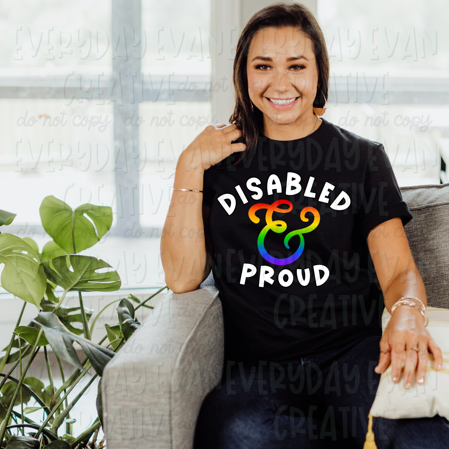 A brunette white woman is sitting on a couch modeling a black t-shirt. The design on the shirt has white block text that says "disabled" curved over a gradient rainbow script ampersand sign. Underneath is curved white block lettering that says "proud."