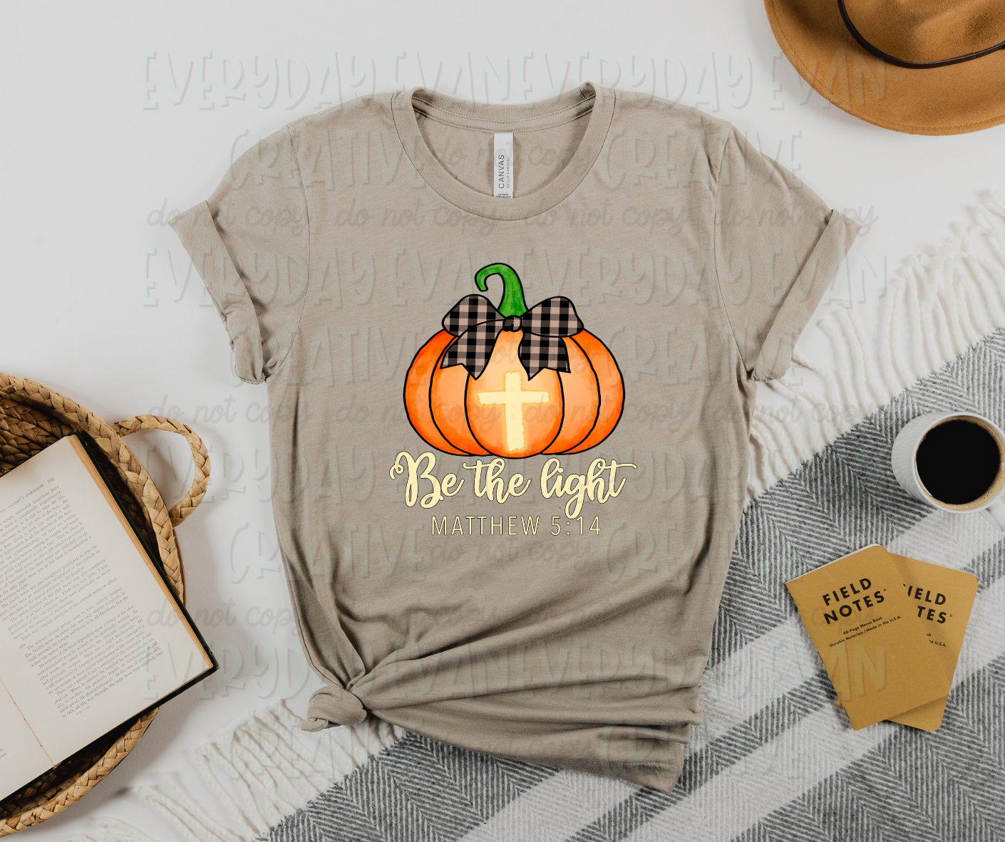 Image displays a t-shirt surrounded by miscellaneous desk items. The shirt's image is an orange pumpkin with a black and tan checkered bow on top of it. The pumpkin has a gold cross in its center. Under the pumpkin there is script text that reads "be the light" and under that there is more text that reads "Matthew 5:14" in the same color as the cross.