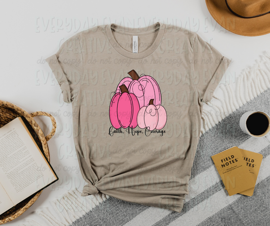Image displays a t-shirt surrounded by miscellaneous desk items. The shirt's image includes three differently sized pink pumpkins. Under the pumpkins, black script text reads "faith, hope, courage"