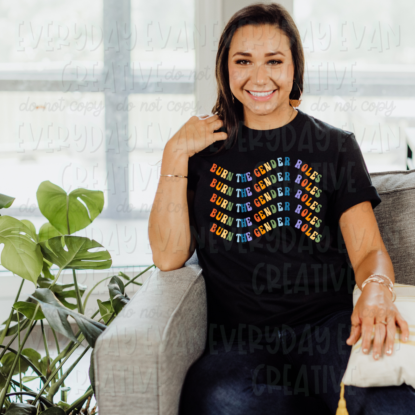 A brunette white woman sits on a couch modeling a black t-shirt. The design on the shirt says "burn the gender roles" in a rainbow wavy retro font. The phrase is stacked and repeated five times.