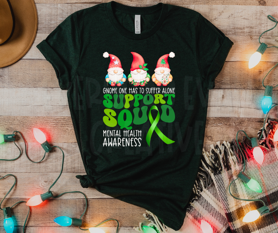 a heather emerald t-shirt with a design that has three Christmas gnomes at the top. underneath it says "gnome one has to suffer alone" then there is a green retro typographic that says "support squad" the "a" in squad is a green awareness ribbon. in the bottom corner it says "mental health awareness"