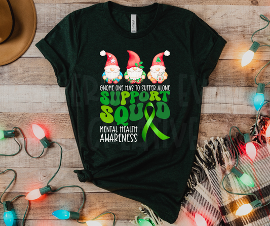 a heather emerald t-shirt with a design that has three Christmas gnomes at the top. underneath it says "gnome one has to suffer alone" then there is a green retro typographic that says "support squad" the "a" in squad is a green awareness ribbon. in the bottom corner it says "mental health awareness"
