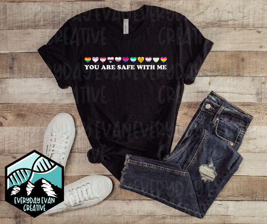 the image is a flat lay of a black t shirt with a pair of jeans next to it. the background a rustic wood. the design on the shirt is a row of LGBTQIA+ themed hearts that says "you are safe with me" in white block letters underneath. the design is thin and goes right across the chest. 